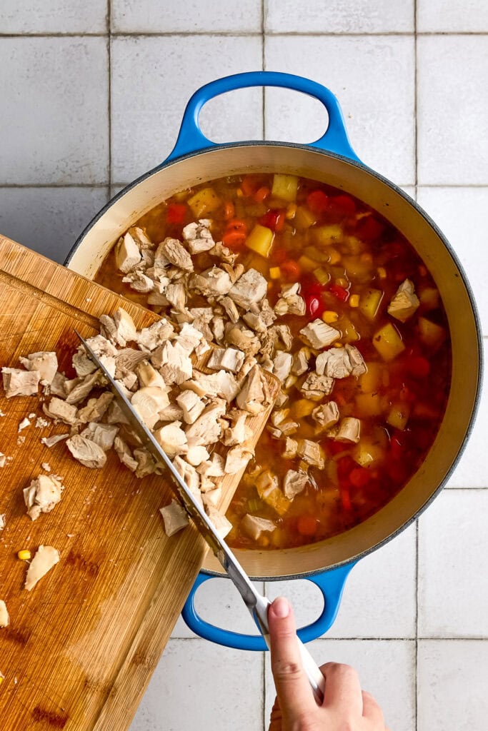Person chopping cooked chicken into a pot of chicken and corn chowder with carrots, potatoes, and broth, on a white tiled surface.