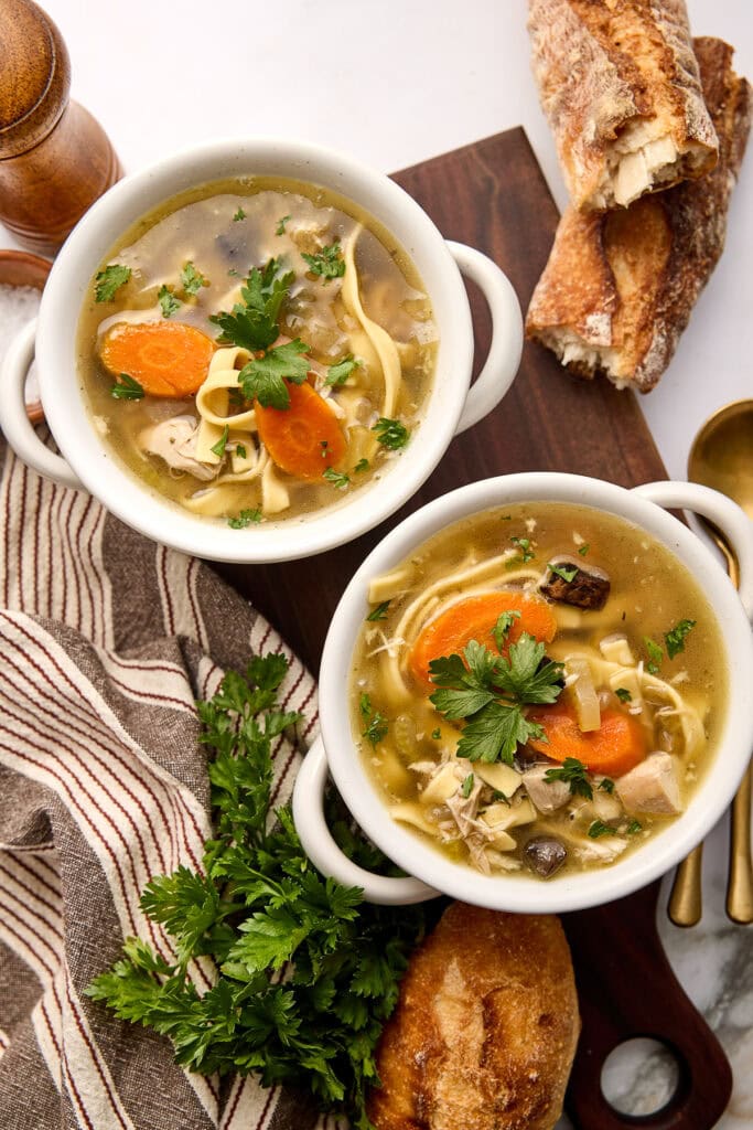 Warm bowls of creamy chicken noodle soup with carrot slices, fresh parsley, and crusty bread on the side.