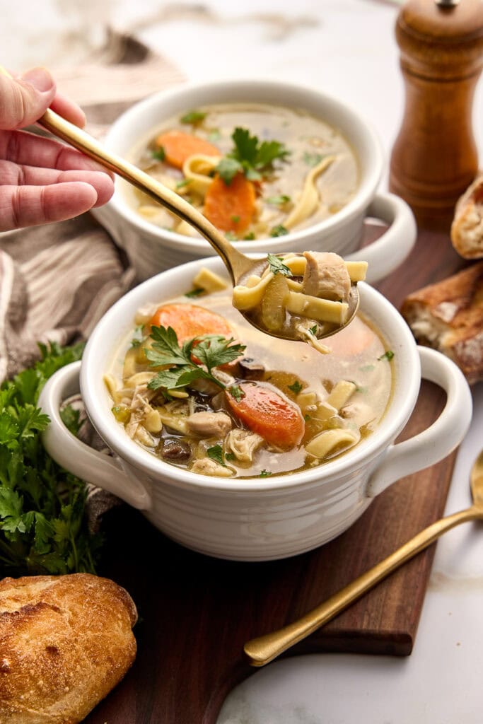Creamy crock pot chicken noodle soup with herbs, served in a white ramekin with a spoon, garnished with parsley and carrot slices, on a wooden board.