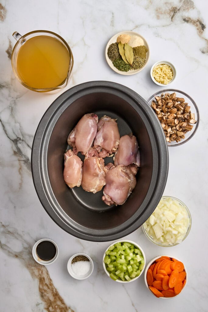 Raw chicken thighs arranged in a slow cooker surrounded by chopped vegetables, spices, and ingredients on a marble surface, ready for slow cooking.
