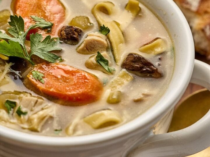 Bowl of creamy chicken noodle soup with carrot slices, herbs, and vegetables in a white mug on a wooden table.