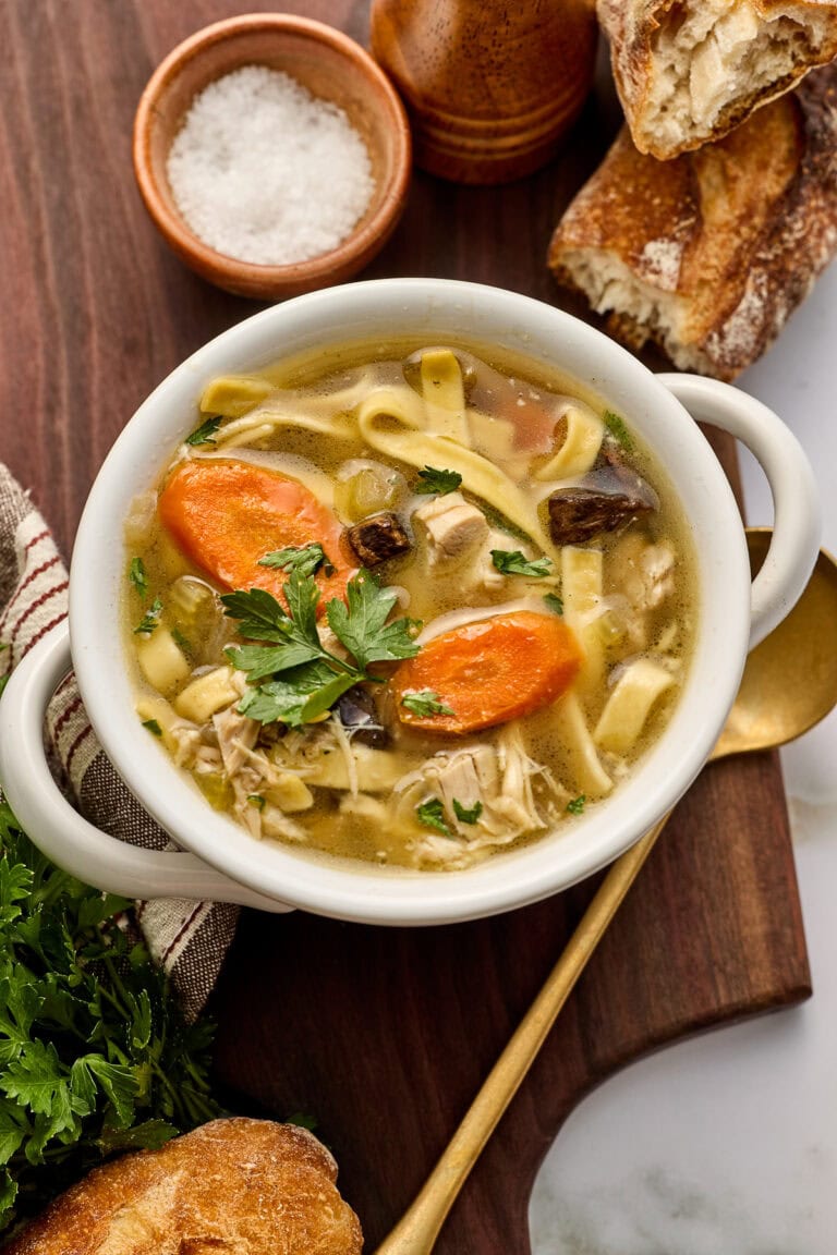 Creamy crock pot chicken noodle soup with carrots, noodles, and herbs served in a white bowl, garnished with parsley, on a wooden board with bread and salt nearby.