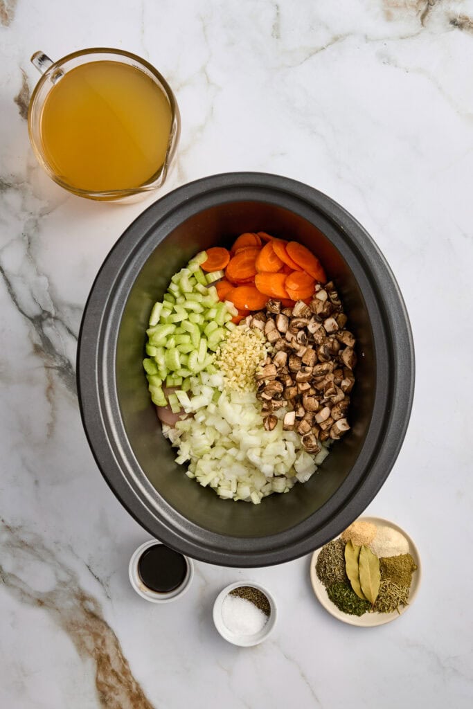 Slow cooker meal bowl with chopped celery, carrots, onions, mushrooms, and seasonings next to a cup of broth.
