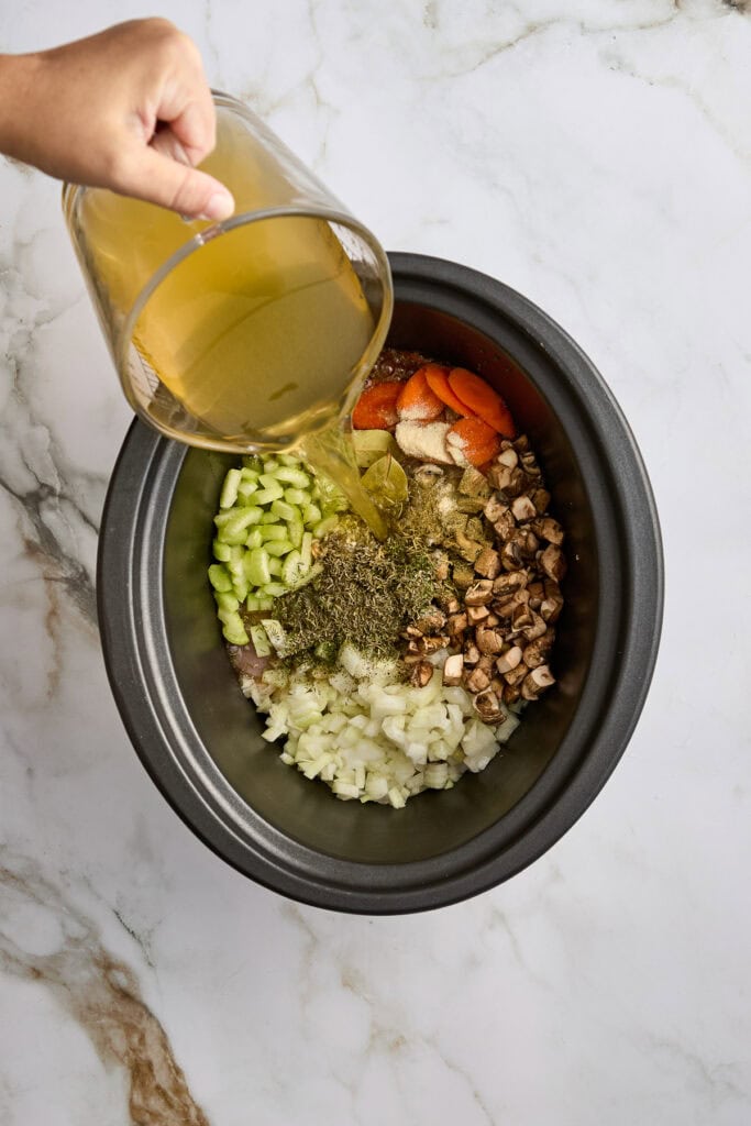 Person pouring broth into a slow cooker filled with diced vegetables and herbs for a hearty veggie stew.