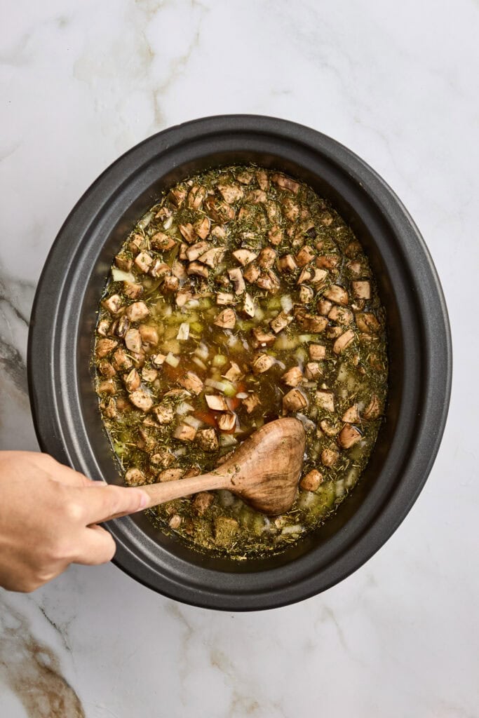 A simmering slow cooker filled with diced vegetables and herbs in a savory broth, being stirred by a wooden spoon.