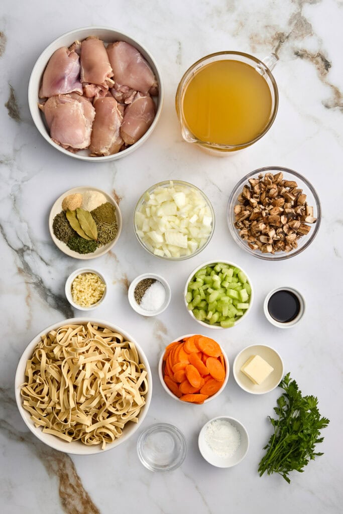 Fresh ingredients for a savory chicken pasta dish: chicken, pasta, celery, onions, carrots, herbs, chopped greens, and a glossy sauce, arranged on a marble surface.