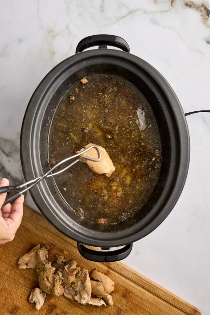 Slow cooker pot with broth and shredded chicken resting on cutting board, hand using tongs to lift a piece; steam and herbs visible in the dark broth.