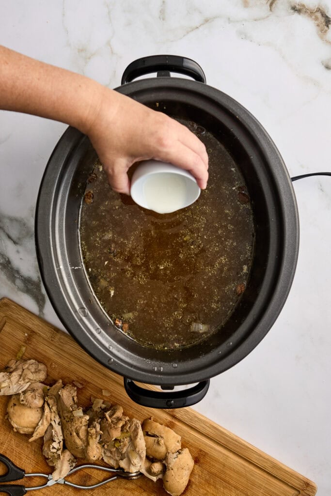 Person pours milk into a simmering Crock-Pot chicken noodle soup, sitting on a marble countertop with a cutting board of crumbled chicken nearby.