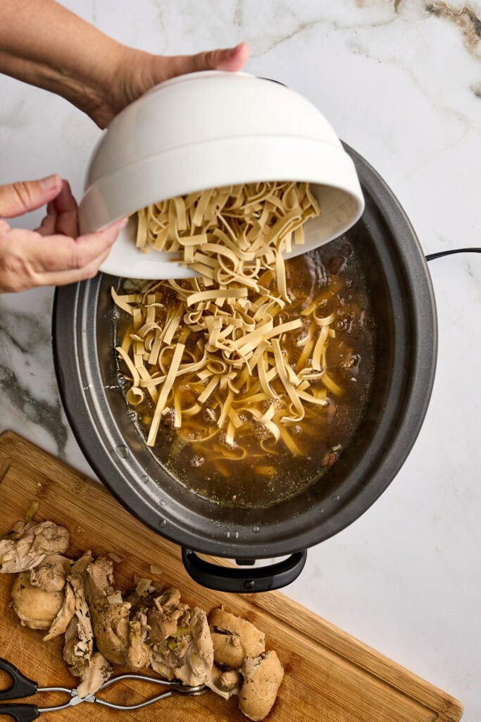 Person pouring shredded noodles into a bubbling slow cooker with pieces of bread on a wooden board nearby.