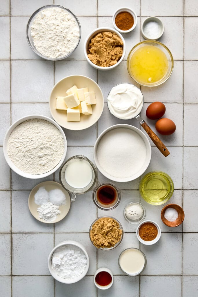 Assorted baking ingredients including flour, butter, eggs, sugar, vanilla, and spices arranged on a white tiled surface.