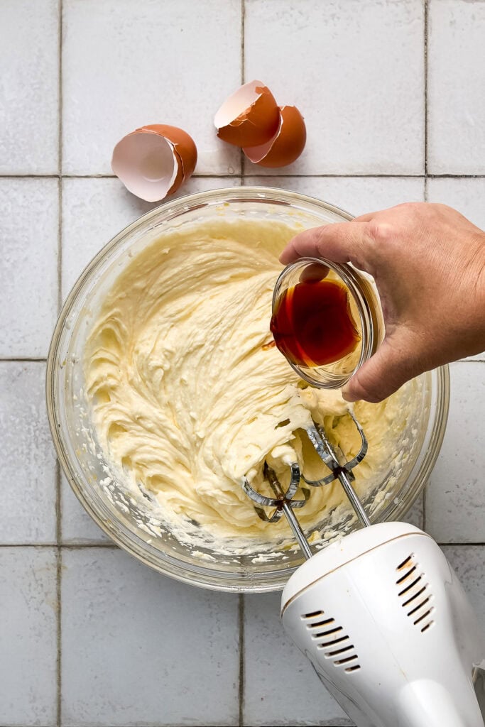 Hand pouring vanilla extract into cinnamon coffee cake batter in a glass mixing bowl, with eggshells nearby on a tiled surface.