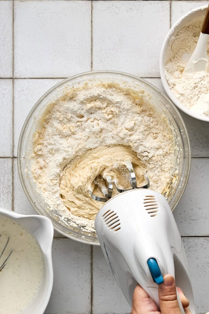 Person mixing cake batter with an electric hand mixer in a glass bowl, surrounded by bowls of flour and milk on a white tiled surface.
