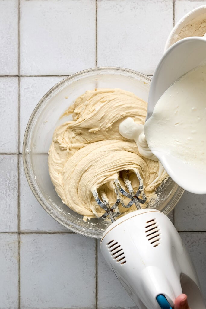 Close-up of cake batter being mixed with a hand mixer, with milk being poured into the bowl, on a white tiled surface.