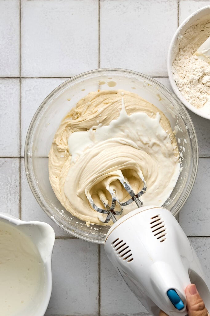 Close-up of cake batter being mixed with an electric hand mixer in a glass bowl, with flour and other ingredients nearby on a tiled surface.