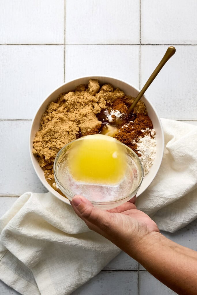 Baking ingredients including brown sugar, cinnamon, and butter in a bowl with a hand holding a small glass bowl of melted butter.