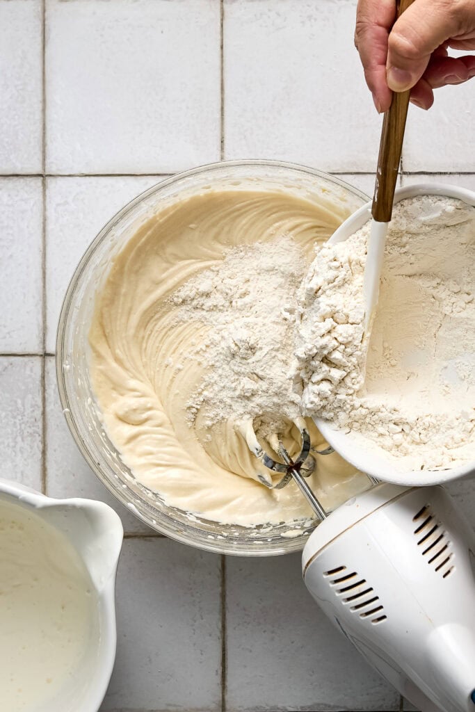 Close-up of a hand mixing cake batter with flour in a glass bowl, with a hand mixer and a small bowl of flour on a white tiled surface.