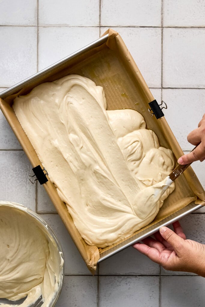 Cinnamon coffee cake batter being spread in a baking pan with a spatula, ready to bake, on a tiled surface.