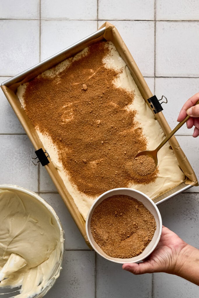 Person sprinkling cinnamon sugar mixture over unbaked cinnamon coffee cake in a baking tray, with a bowl of cinnamon sugar and a spatula nearby.