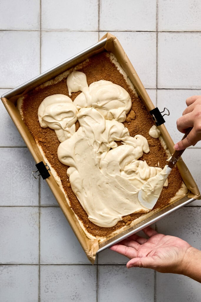Cinnamon coffee cake batter being spread in a baking pan with a spatula, ready to bake, on a tiled surface.