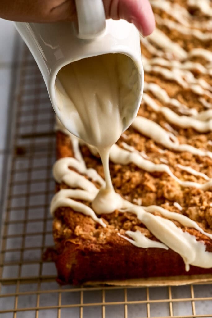 Close-up of creamy white glaze being poured over freshly baked cinnamon crumb coffee cake on a cooling rack.