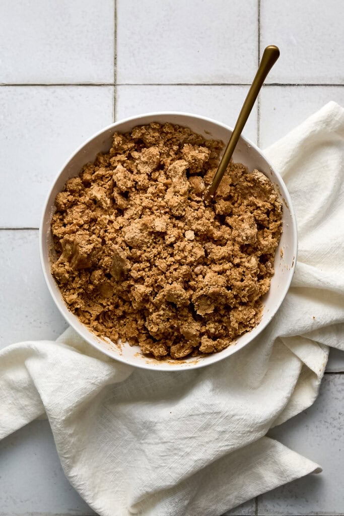 Close-up of a bowl of crumb topping for cinnamon coffee cake with a spoon on a white cloth napkin.