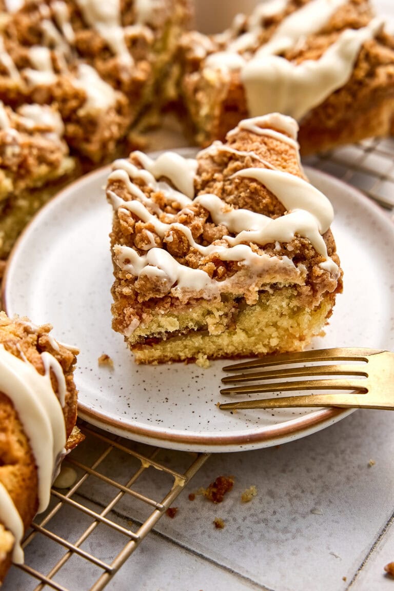 Close-up of a moist cinnamon coffee cake slice with white icing drizzle on a white plate, showcasing a golden crust and crumb topping.