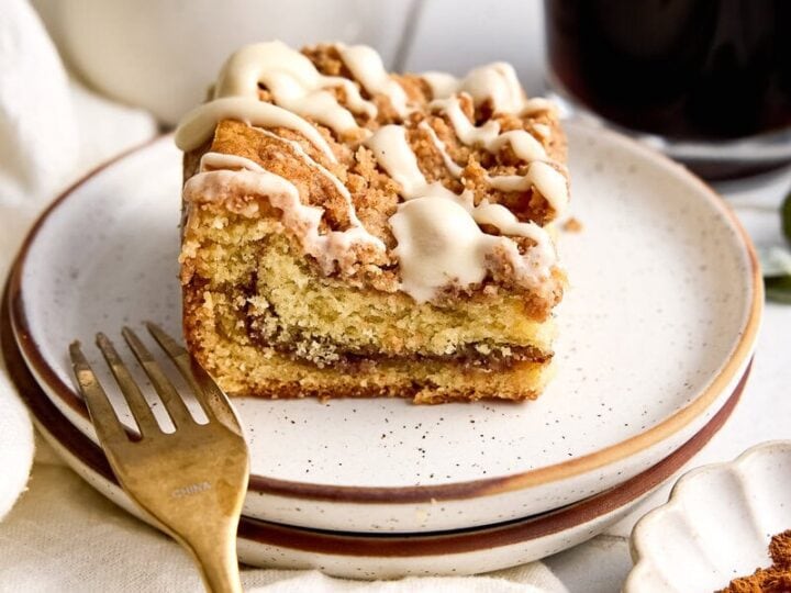 A slice of cinnamon coffee cake with icing on a white plate, served with a glass of dark coffee and a fork, on a rustic table setting.