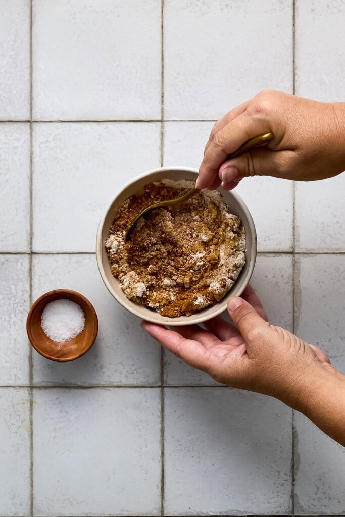 Hands mixing baking ingredients in a bowl on a white tiled surface, with a small bowl of salt nearby, for cinnamon coffee cake