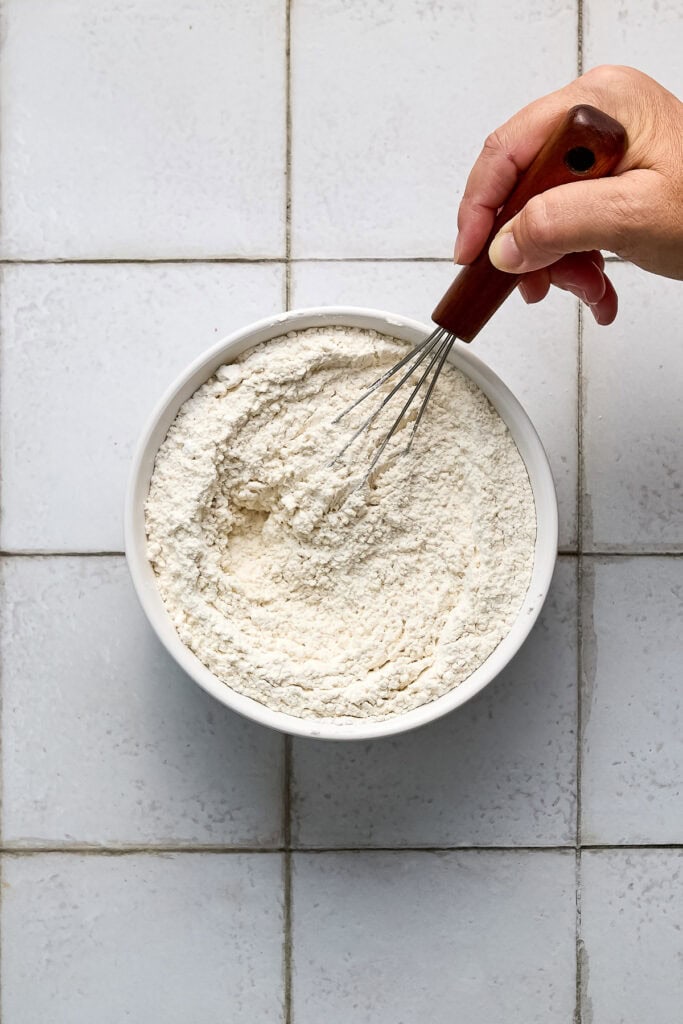 Close-up of a hand holding a whisk over a bowl of flour on a tiled surface, ready for baking.