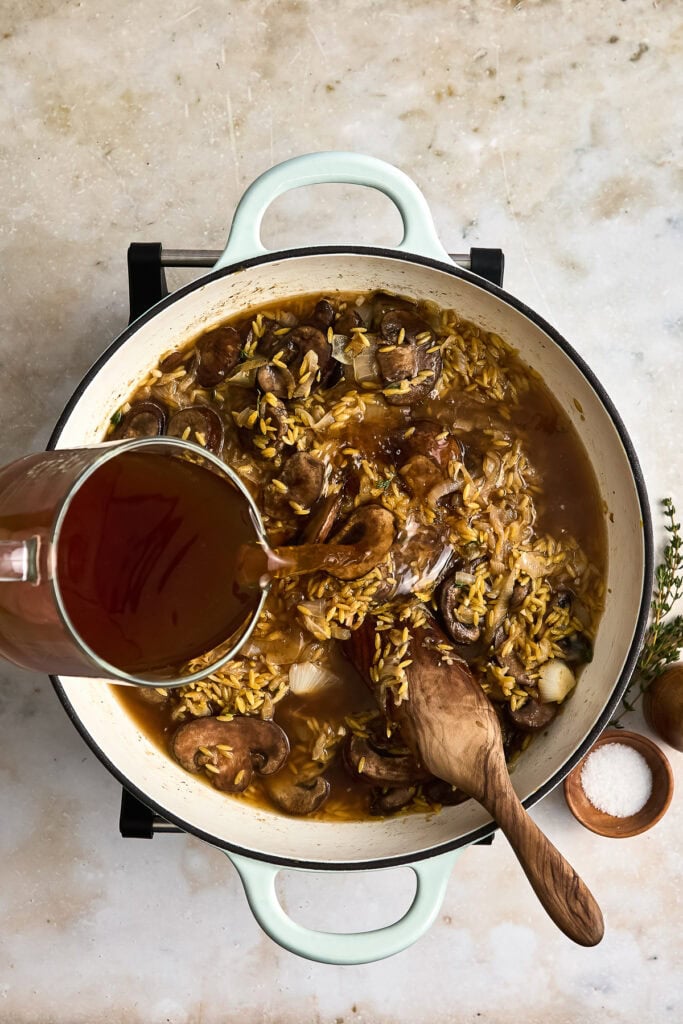A pot of mushroom and orzo being prepared with broth poured in, featuring fresh mushrooms and herbs on a rustic surface.