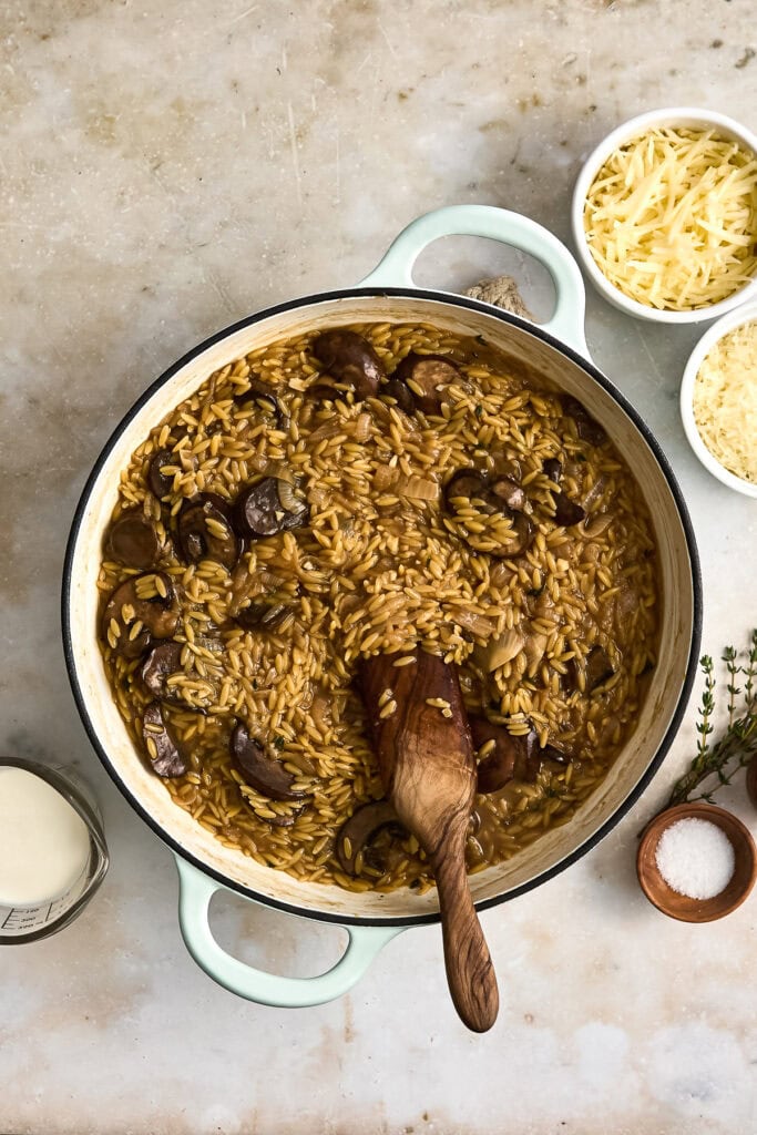 Creamy mushroom orzo with sliced mushrooms and herbs in a white cooking pot, served with grated cheese and side ingredients on a light surface.