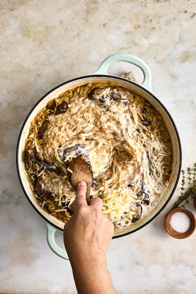 A hand stirring a creamy onion and mushroom pasta dish in a large pot, with ingredients like shredded cheese and herbs nearby.