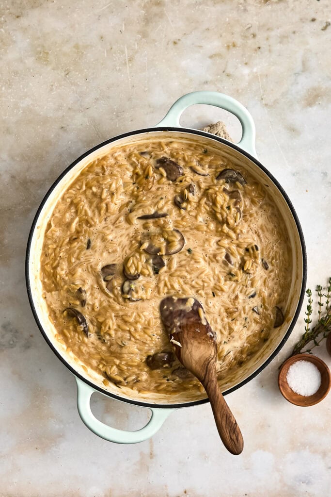 A creamy mushroom French onion pasta in a light green skillet with a wooden spoon, served on a rustic surface with salt and herbs nearby.