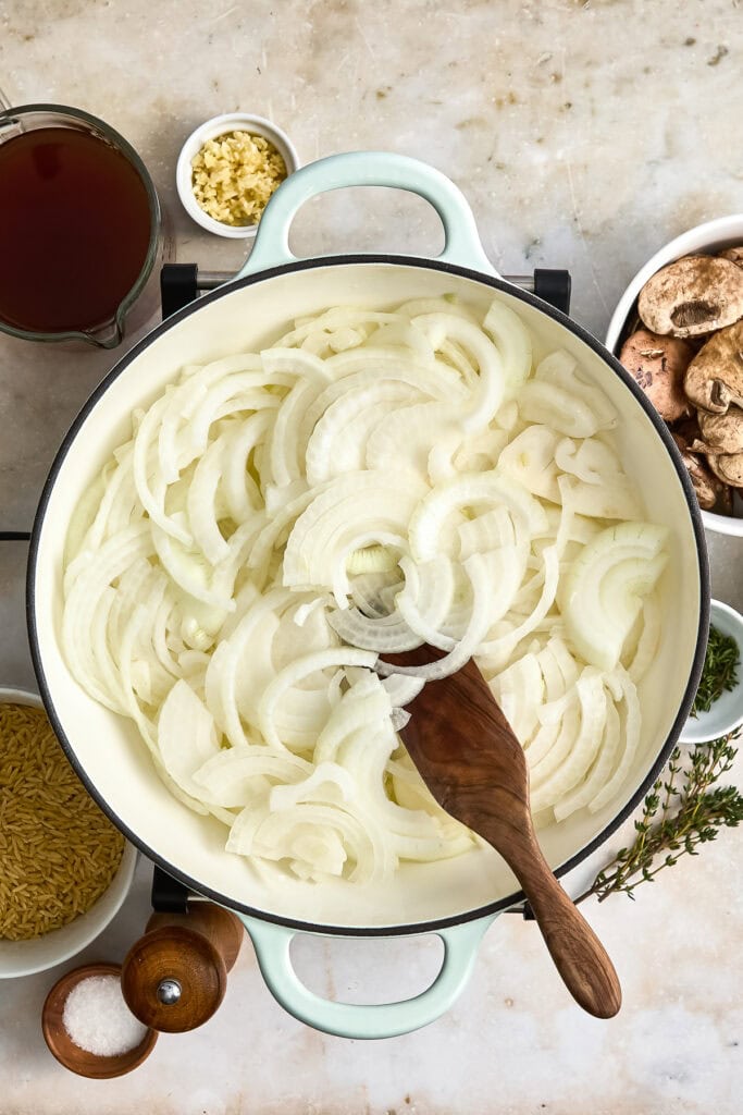 A pot of sliced onions cooking surrounded by ingredients like garlic, mushrooms, and spices for a flavorful onion soup.