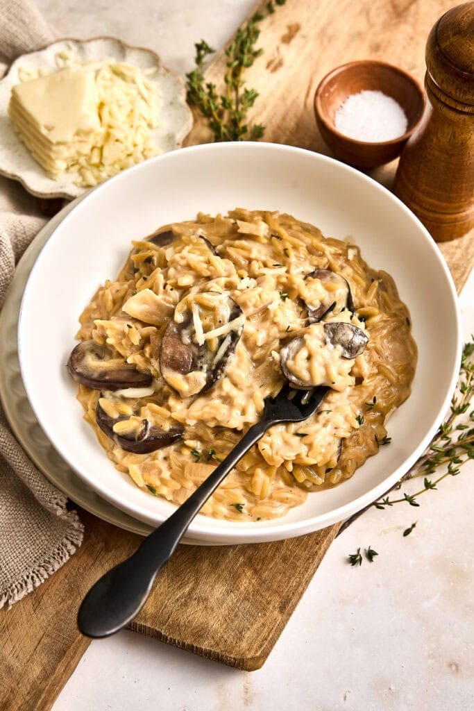 A bowl of creamy French onion and mushroom orzo with sliced mushrooms and grated cheese, served on a wooden board with butter, herbs, and seasoning nearby.