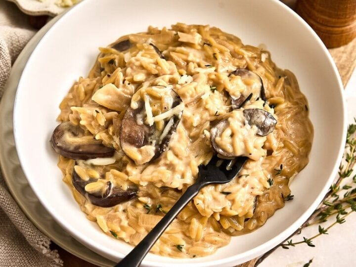 A bowl of creamy French onion pasta with sliced mushrooms and grated cheese, served on a wooden board with butter, herbs, and seasoning nearby.