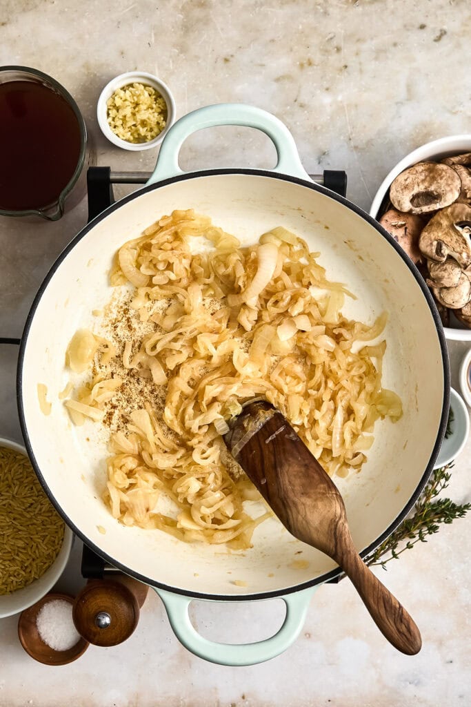 Close-up of caramelized onions cooking in a white pot with garlic, herbs, and side ingredients on a kitchen countertop.