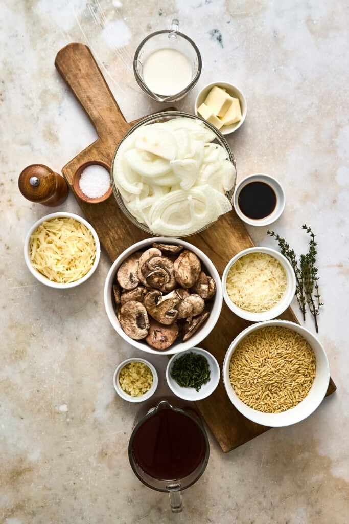 Assorted ingredients for French onion mushroom pasta including cream, mushrooms, cheese, and spices on a wooden board.