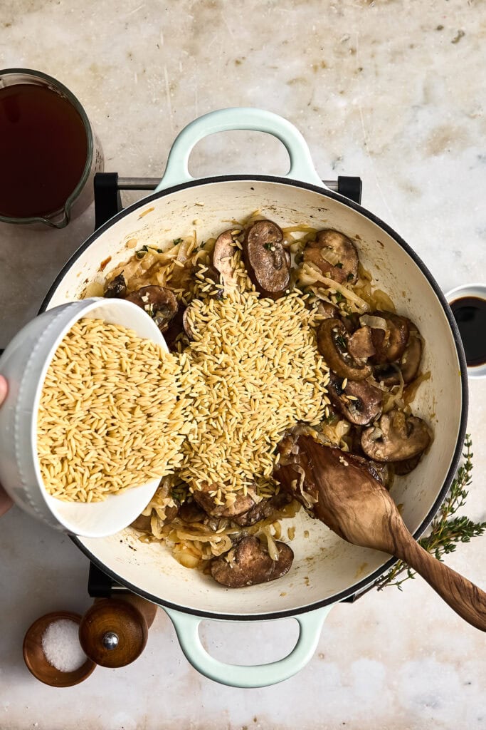Step-by-step image of adding orzo pasta to a pan with cooked mushrooms and onions for a French onion pasta recipe